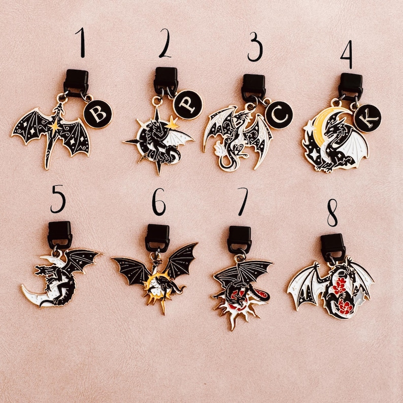 May include: Collection of eight dragon-themed charms with black clasps. Each charm features a different dragon design in black, white, and gold, with some including red accents. Each charm has a letter on a small circular charm.
