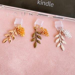 Könnte beinhalten: Drei dekorative Telefonanhänger mit Blatt- und Tannenzapfenanhängern. Ein Anhänger hat ein goldenes Blatt und einen goldenen Tannenzapfen, ein anderer ein bronzefarbenes Blatt und einen goldenen Tannenzapfen, und der dritte ein silbernes Blatt und einen silbernen Tannenzapfen.