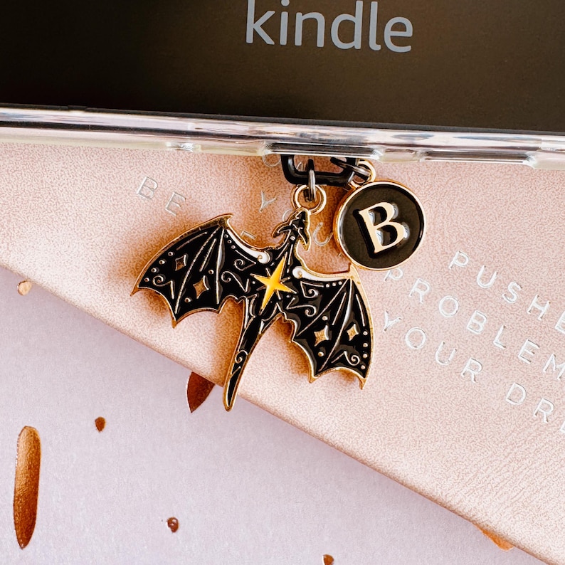 May include: A black and gold dragon charm with a yellow star accent and a letter "B" charm. The dragon charm is attached to a clear phone case, resting on a pink surface with the word "kindle" visible.
