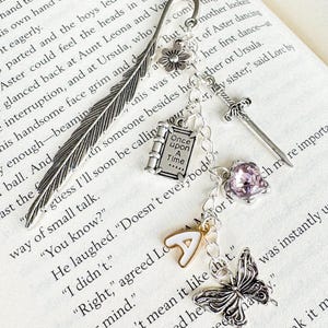 May include: A silver bookmark with a feather design, a flower, a book charm with the text "Once Upon A Time", a sword, a purple gem, a letter "A" charm, and a butterfly. The bookmark is on the pages of a book.