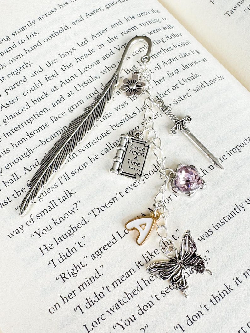 May include: A silver bookmark with a feather design, a flower, a book charm with the text "Once Upon A Time", a sword, a purple gem, a letter "A" charm, and a butterfly. The bookmark is on the pages of a book.
