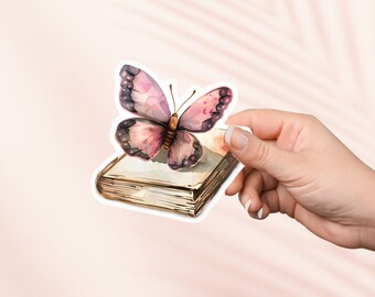 Puede incluir: Una ilustración de acuarela de una mariposa rosa y morada posada sobre un libro cerrado con una estética vintage.