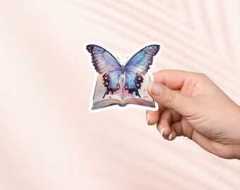 Puede incluir: Una pegatina con una mariposa azul y morada con alas iridiscentes posada sobre un libro abierto. El libro es blanco con detalles dorados y tiene un aspecto vintage.