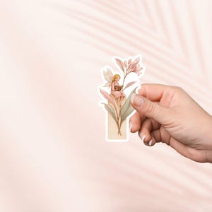 May include: A sticker featuring a watercolor illustration of a fairy reading a book while sitting on a flower. The fairy has blonde hair and wears a pink dress. The sticker has a light pink, beige, and green color palette.