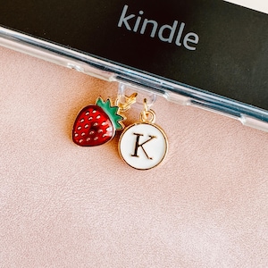 May include: A clear phone case with a gold charm featuring a red strawberry and a white circular charm with the letter "K". The word "kindle" is visible on the phone.