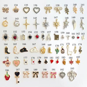 May include: A collection of small, decorative charms in various shapes and designs. The charms include bows, hearts, crosses, stars, and other symbols. They are made of metal and enamel, with a variety of colors and finishes.