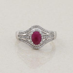 May include: A silver-toned ring featuring a central oval-cut deep red gemstone. The stone is surrounded by a halo of small clear stones, with additional clear stones set along the split-shank band that curves around the center setting.