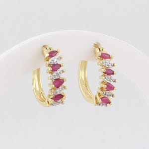 May include: A pair of gold hoop earrings featuring a row of alternating marquise-cut red rubies and round white diamonds. The earrings have a hinged closure mechanism and are displayed on a curved white stand, showcasing the gemstone arrangement.