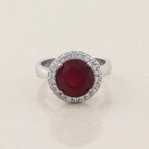 May include: A silver-toned ring featuring a large, round, deep red center stone. The central red stone is surrounded by a halo of small, clear, sparkling stones set in a circular frame. The band of the ring is smooth, polished, and metallic.