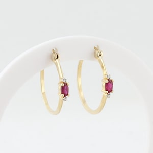 May include: A pair of gold hoop earrings, each with an oval ruby and two small diamonds. The earrings are presented on a white stand against a white backdrop. The ruby is a deep red colour.