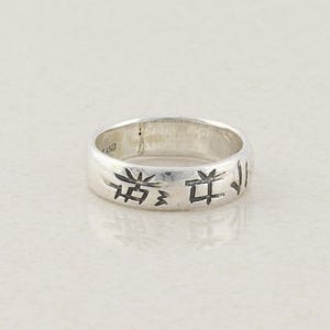 Puede incluir: Un anillo de plata con símbolos grabados. El anillo tiene una banda con una serie de símbolos grabados alrededor de la circunferencia. El anillo está sobre un fondo claro.