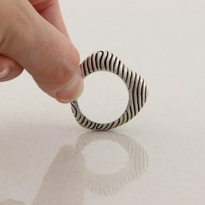 May include: A silver ring with a unique design. The ring has a rounded square shape with vertical, wavy lines carved into the metal. The ring is held up by a hand against a white background.
