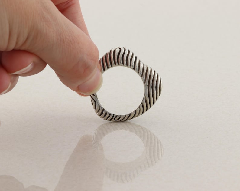 May include: A silver ring with a unique design. The ring has a rounded square shape with vertical, wavy lines carved into the metal. The ring is held up by a hand against a white background.