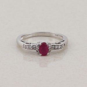 May include: A silver-toned ring featuring a central oval-cut red gemstone held in a prong setting. The band is accented with small, clear, round-cut stones embedded along the shoulders on both sides of the center stone, creating a symmetrical design.