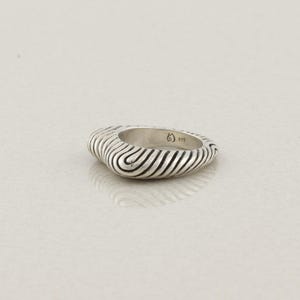 May include: A silver ring with a textured, ridged design. The ring has a rounded, oval shape and a polished finish. The ridges create a wave-like pattern around the band. The ring is likely made of sterling silver.