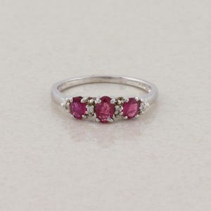 May include: A silver ring featuring three oval-cut, vibrant red gemstones, likely rubies, set in a row. Small, clear accent stones are interspersed between the larger gems. The ring has a simple, elegant design.