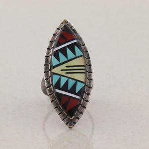 May include: A silver ring with a long, oval face. The face is inlaid with a geometric pattern of black, red, turquoise, and cream-colored stones. The ring has a textured border and a simple band.