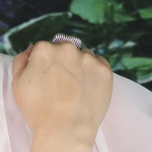 May include: A silver-toned ring with a ribbed, rectangular design. The ring is worn on a hand, with the ribbed texture creating a series of parallel lines across the top. The background is a blurred mix of green and white.