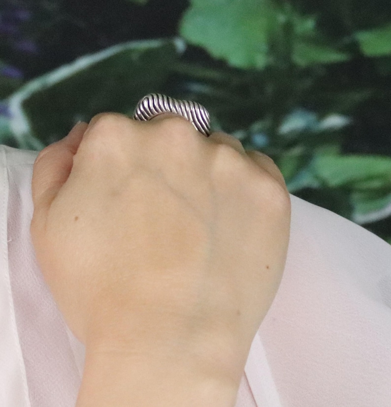 May include: A silver-toned ring with a ribbed, rectangular design. The ring is worn on a hand, with the ribbed texture creating a series of parallel lines across the top. The background is a blurred mix of green and white.