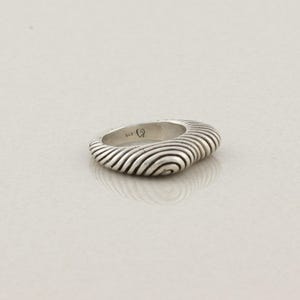 May include: A silver ring with a unique design. The ring has a square shape with rounded edges and a textured surface featuring concentric, wave-like lines. The ring is set against a plain, light background.