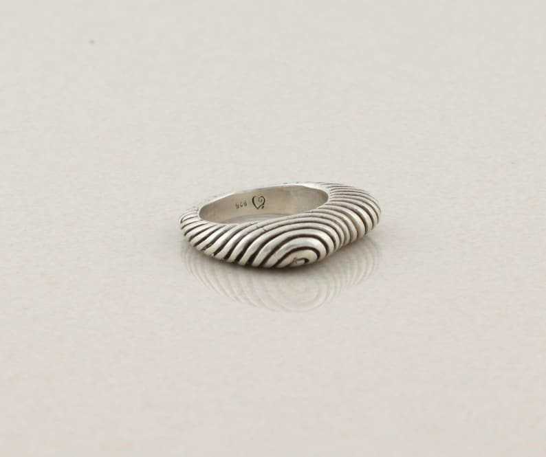 May include: A silver ring with a unique design. The ring has a square shape with rounded edges and a textured surface featuring concentric, wave-like lines. The ring is set against a plain, light background.