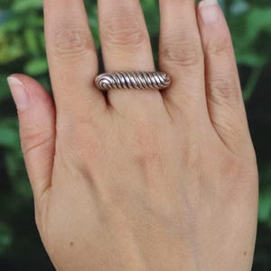 May include: A silver ring with a spiral design. The ring is worn on a finger and has a textured, ridged appearance. The ring's design resembles a tightly wound coil or spring. The ring is a simple, yet elegant piece of jewelry.