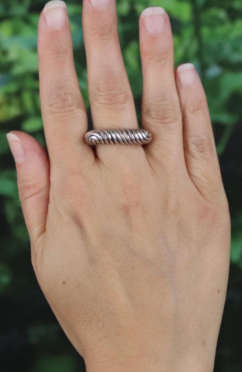 May include: A silver ring with a spiral design. The ring is worn on a finger and has a textured, ridged appearance. The ring's design resembles a tightly wound coil or spring. The ring is a simple, yet elegant piece of jewelry.