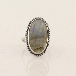 May include: An oval-shaped labradorite gemstone ring set in a polished silver band. The stone features a smooth surface with iridescent blue and grey tones, surrounded by a decorative twisted rope-style metal border frame.
