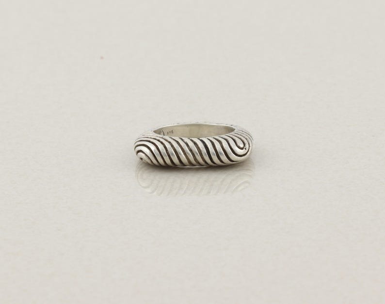 May include: A silver ring with a textured, ridged design. The ring's surface features a series of curved, parallel lines, creating a wave-like pattern. The ring is set against a plain, light-colored background.