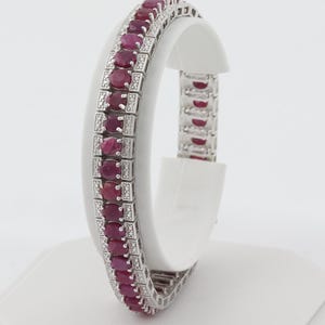 May include: A silver-toned bracelet featuring a central row of oval-cut deep red rubies. The rubies are flanked by two parallel rows of small, clear, round-cut stones set in a rectangular metal frame, creating a structured and repeating pattern.