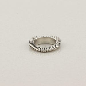 May include: A silver ring with a textured, ridged design. The ring's surface features a series of curved lines, creating a unique pattern. The ring is a simple band style, suitable for everyday wear.