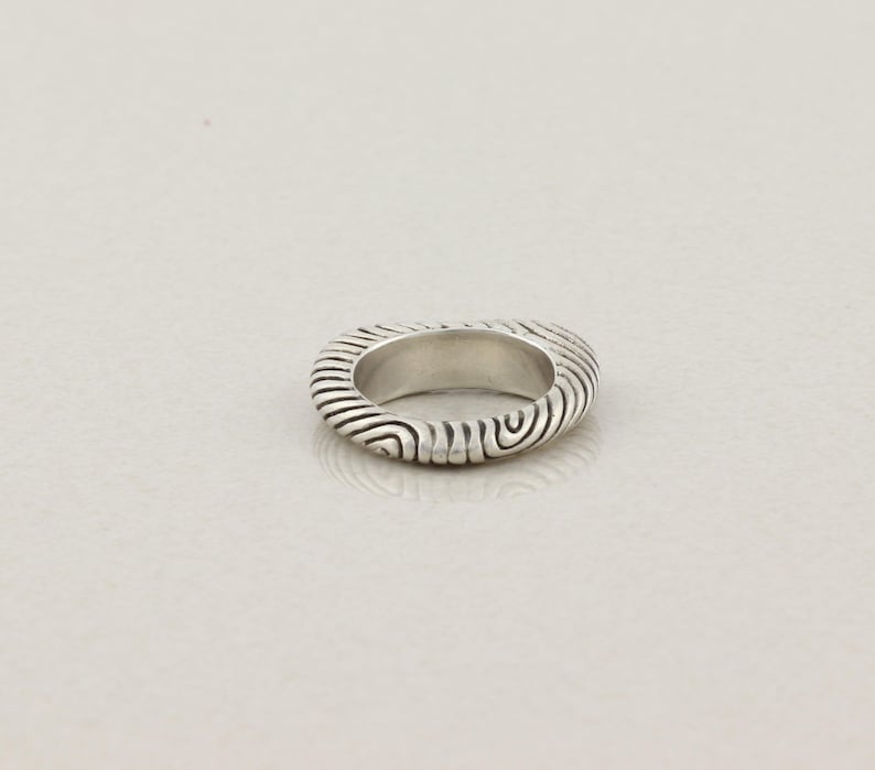 May include: A silver ring with a textured, ridged design. The ring's surface features a series of curved lines, creating a unique pattern. The ring is a simple band style, suitable for everyday wear.