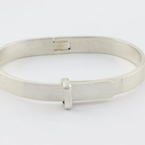 Peut inclure: Un bracelet jonc en métal argenté à la surface lisse et plate. Le bracelet présente un mécanisme de fermoir à charnière sur le dessus et un détail de boucle coulissante rectangulaire positionné à l'avant, formant un cercle uniforme.