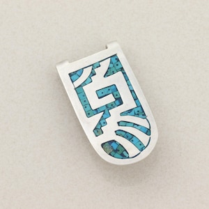 Könnte beinhalten: Ein silberfarbener Geldclip mit türkisfarbener Einlage in einem geometrischen Design. Der Clip hat eine rechteckige Form mit abgerundeten Kanten und einem Klappdeckel. Das Türkis ist ein Mosaik aus kleinen, unregelmäßigen Teilen.