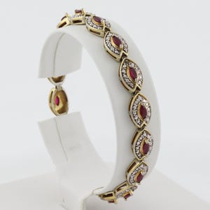 May include: A gold-toned bracelet featuring a series of marquise-shaped links. Each link contains a central oval-cut red gemstone surrounded by a border of small, clear, round-cut stones set in a pavé style, creating a repeating floral pattern.