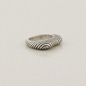 May include: A silver ring with a unique, textured design. The ring features a series of concentric, curved lines that create a wave-like pattern. The ring is a solid, rounded shape, and the metal has a polished, reflective surface.