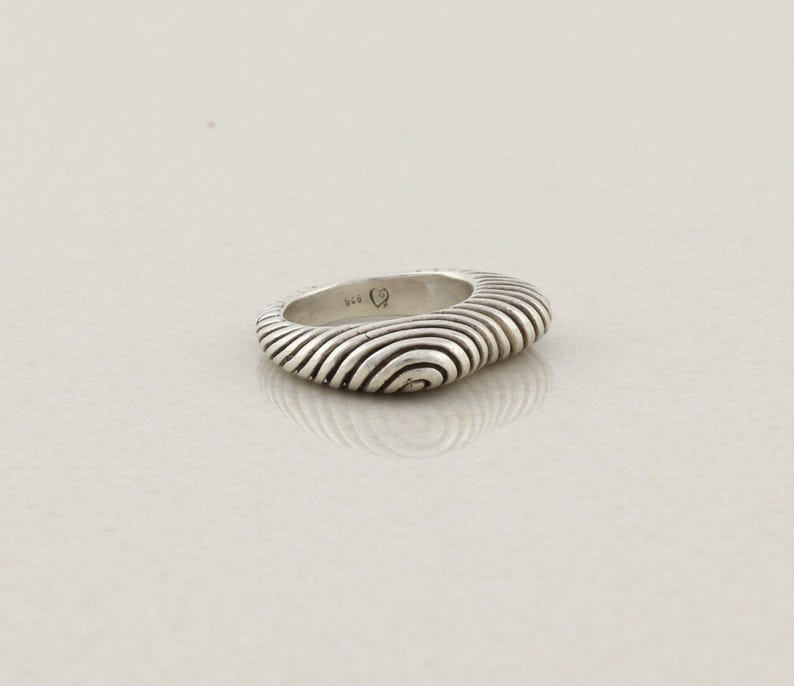 May include: A silver ring with a unique, textured design. The ring features a series of concentric, curved lines that create a wave-like pattern. The ring is a solid, rounded shape, and the metal has a polished, reflective surface.