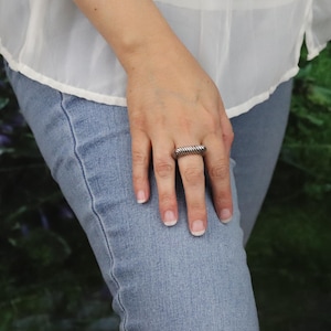 May include: A silver-toned ring with a textured, ridged design. The ring is rectangular and worn on a finger. The person is wearing a white blouse and blue jeans.