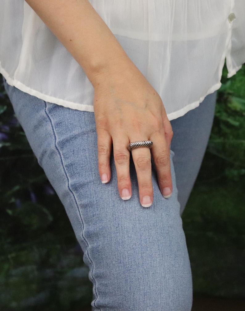 May include: A silver-toned ring with a textured, ridged design. The ring is rectangular and worn on a finger. The person is wearing a white blouse and blue jeans.