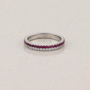 May include: A thin silver-toned metal band ring featuring two parallel rows of small, round-cut stones. The top row consists of vibrant, deep pink-red stones, while the bottom row is composed of clear, sparkling white stones set in a continuous line.