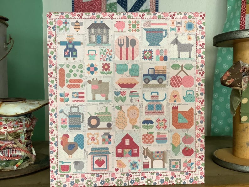 May include: A rectangular quilt featuring a grid of small square blocks with various motifs including a house, farm animals, a truck, a barn, fruits, vegetables, and kitchen utensils, all framed by a floral border in pink, blue, and green.