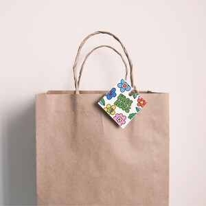 Peut inclure: Un sac cadeau en papier brun avec des poignées torsadées. Une étiquette blanche en forme de losange avec des fleurs colorées et les mots "FREE HUGS" est suspendue au sac. L'étiquette est attachée avec de la ficelle.
