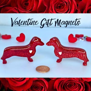 May include: Two red glitter dachshund shaped Valentine's Day magnets with white hearts. The magnets are next to a penny for size comparison. Valentine Gift Magnets.