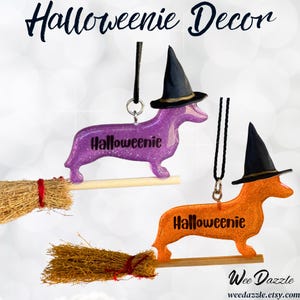 May include: Two hanging ornaments shaped like dachshunds, one purple and one orange, both with glittery finishes. Each dog wears a black witch hat and is attached to a miniature broomstick. The text "Halloweenie" is printed on each dog.