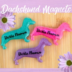 May include: Three dachshund-shaped magnets with glitter finish. Each magnet is a different color: teal, pink, and purple. The magnets are on a wooden surface and each has the text "Doxie Mama" written on it.