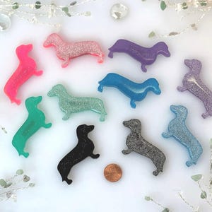 May include: A collection of ten small dachshund-shaped resin ornaments in various colors including pink, purple, blue, green, black, and silver glitter. A copper-colored penny is placed near the center for scale, showing the size of the figures.