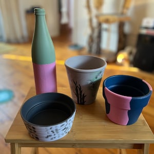 Könnte beinhalten: Vier bemalte Keramik-Blumentöpfe in verschiedenen Farben und Designs. Eine grüne und rosa Flasche ist ebenfalls enthalten. Die Töpfe stehen auf einem Holztisch.