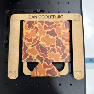 May include: A brown wooden jig with the text "CAN COOLER JIG" printed on it. A camouflage patterned can cooler is placed inside the jig.