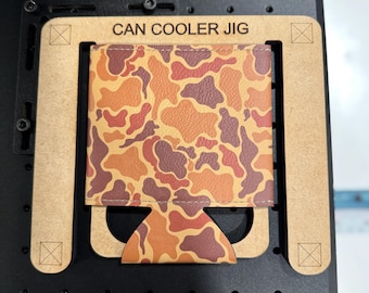 May include: A brown wooden jig with the text "CAN COOLER JIG" printed on it. A camouflage patterned can cooler is placed inside the jig.