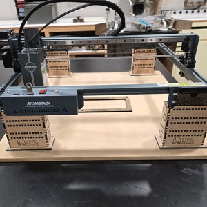 Op de afbeelding: Een grijze en zwarte lasersnijder met een houten basis. De lasersnijder staat op een houten tafel. De lasersnijder heeft een rode knop aan de voorkant. De lasersnijder is gelabeld met "ATOMSTACK  DIY LASER ENGRAVER STUDIO".